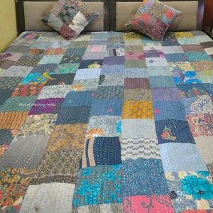 Op de afbeelding: Een patchworkdeken met een blauwe achtergrond en verschillende kleurrijke patronen. De deken is gemaakt van vierkanten van verschillende stoffen, sommige met bloemenpatronen, sommige met geometrische patronen en sommige met paisleypatronen. De deken ligt op een bed met twee kussens, een met een bloemenpatroon en een met een geometrisch patroon.