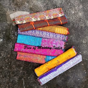 May include: A collection of folded fabric textiles featuring various patterns and colors including orange, pink, blue, purple, and floral prints. The stack includes diverse textures and designs with the text Pics of Mastery India visible.
