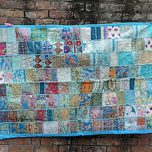 May include: A rectangular patchwork wall hanging featuring a grid of various fabric squares in shades of blue, turquoise, gold, and pink. The textile displays diverse floral, geometric, and metallic embroidered patterns, all bordered by a solid blue trim.