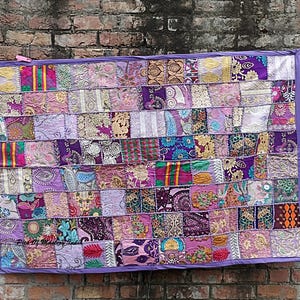 May include: A rectangular wall hanging featuring a patchwork design composed of various fabric squares in shades of purple, pink, gold, and teal. The textile displays diverse floral, paisley, and geometric patterns with embroidered details and a purple border.