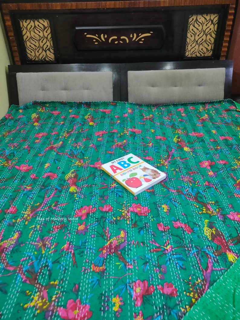 May include: A green kantha quilt with a floral and bird print. The quilt is on a bed with a brown headboard. A children's book titled "My First ABC Board Book" is on top of the quilt.