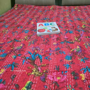 May include: A red kantha quilt with a floral and bird pattern. The quilt is on a bed with a book titled "My First ABC Board Book" on top of it.