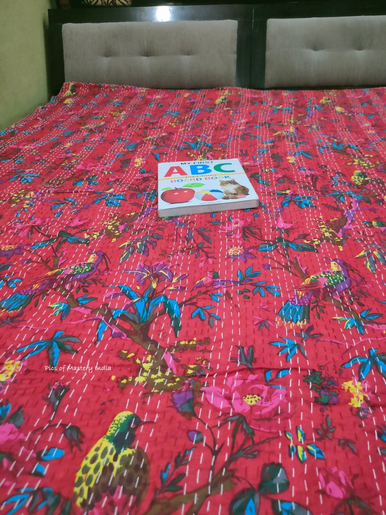 May include: A red kantha quilt with a floral and bird pattern. The quilt is on a bed with a book titled "My First ABC Board Book" on top of it.
