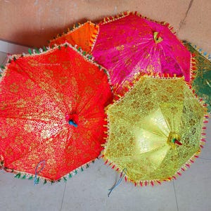 Puede incluir: Una colección de paraguas decorativos coloridos. Los paraguas son de color rojo, rosa, naranja, amarillo y verde. Cada paraguas tiene un patrón floral con detalles dorados y un borde con flecos.