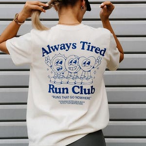 Puede incluir: Camiseta color crema con el texto azul "Always Tired" y "Run Club". La camiseta presenta un gráfico de dibujos animados de tres personajes sonrientes. La frase "RUNS THAT GO NOWHERE" está debajo del nombre del club.