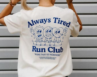 May include: Cream-colored t-shirt with the text "Always Tired" and "Run Club" in blue. The shirt features a cartoon graphic of three smiling characters. The phrase "RUNS THAT GO NOWHERE" is below the club name.