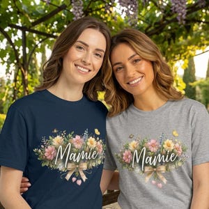May include: Two women wearing t-shirts with a floral design and the word "Mamie". The floral design includes pink, yellow, and blue flowers, with a tan bow. One shirt is navy blue, the other is gray.
