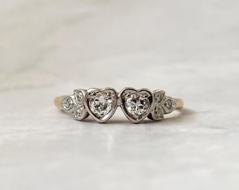 May include: A ring featuring two central heart-shaped settings, each containing a round faceted stone. The band is crafted from a metallic material, with additional small stones set into leaf-shaped accents on either side of the hearts.
