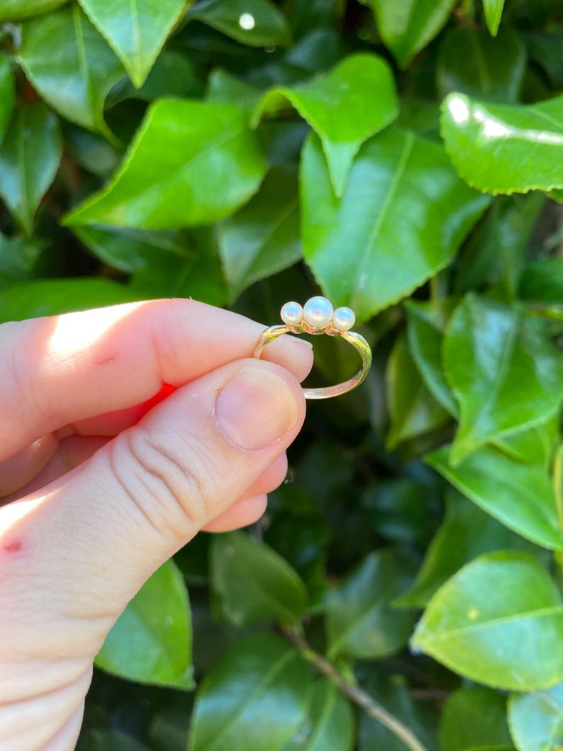 May include: A gold ring featuring three small, round pearls. The ring has a delicate band and the pearls are clustered together. The ring is held against a backdrop of green leaves, creating a natural contrast.
