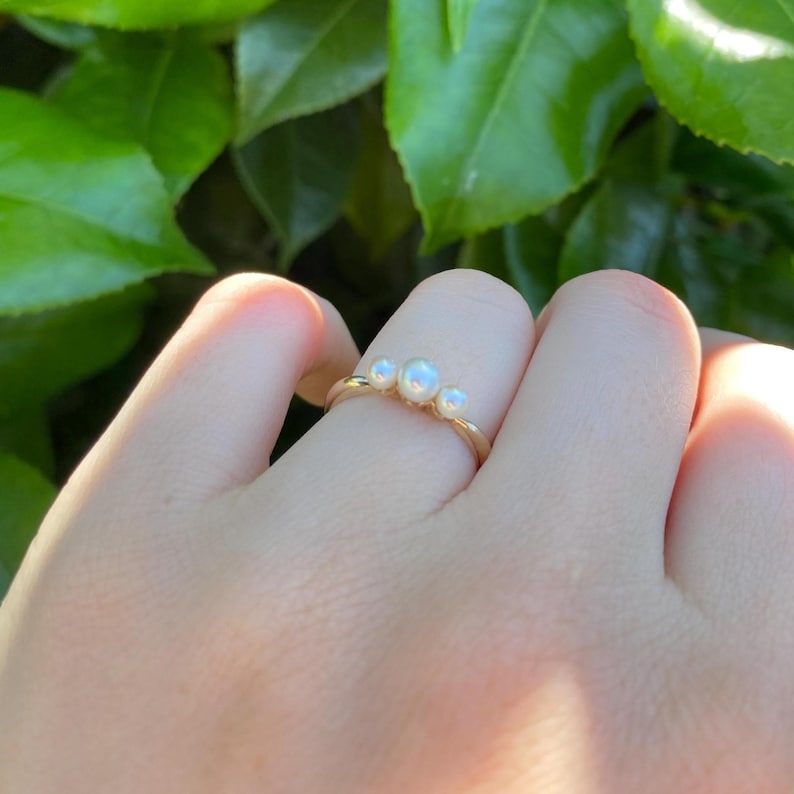 May include: A gold ring featuring three small, round, white pearls. The ring is set on a thin gold band. The background is blurred green foliage.