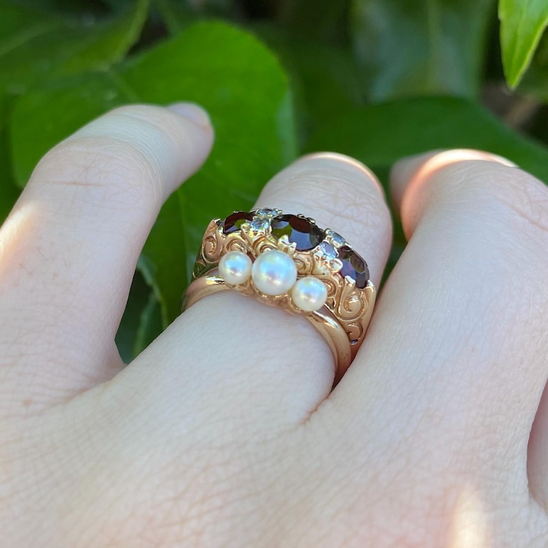 May include: A gold ring with three pearls and several dark red gemstones. The ring has an ornate design with a vintage aesthetic. The ring is on a finger, with green foliage in the background.