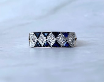May include: A silver ring with a repeating pattern of alternating blue and clear diamond-shaped stones. The ring is set on a white surface, showcasing the intricate design and sparkle of the gemstones.
