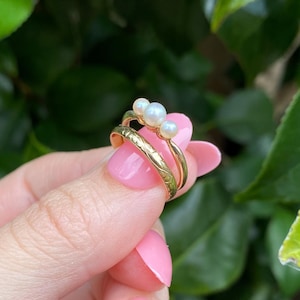 May include: A set of two gold rings, one with a textured band and the other with three small, round pearls. The rings are held up against a background of green leaves.
