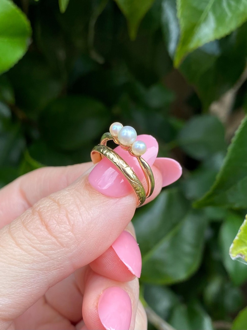 May include: A set of two gold rings, one with a textured band and the other with three small, round pearls. The rings are held up against a background of green leaves.