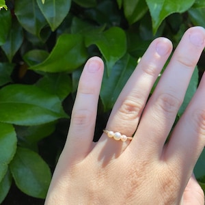 May include: A gold-colored ring with three small, round pearls. The ring is delicate and simple, with the pearls set close together. The ring is shown on a hand, with a background of green leaves.