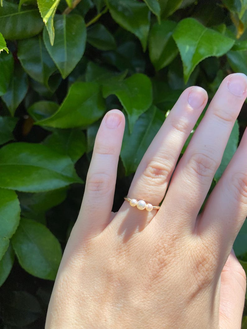 May include: A gold-colored ring with three small, round pearls. The ring is delicate and simple, with the pearls set close together. The ring is shown on a hand, with a background of green leaves.