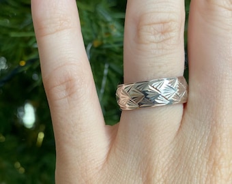 May include: A silver band ring with a geometric pattern. The ring is worn on a person's finger.