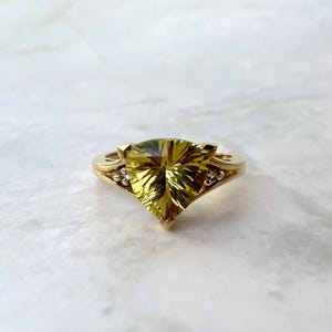 May include: A gold ring featuring a large, faceted, yellow-green gemstone in a unique triangular shape. The ring has a delicate band with small accent stones on each side of the main stone. The setting is a warm gold color.