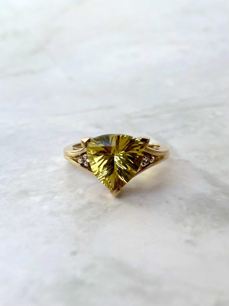 May include: A gold ring featuring a large, faceted, yellow-green gemstone in a unique triangular shape. The ring has a delicate band with small accent stones on each side of the main stone. The setting is a warm gold color.