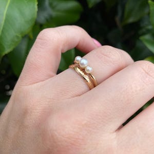 May include: Two gold rings, one with a textured band and the other with three small pearls. The rings are on a hand with a green leafy background.