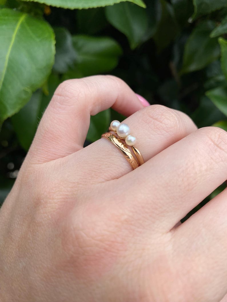 May include: Two gold rings, one with a textured band and the other with three small pearls. The rings are on a hand with a green leafy background.