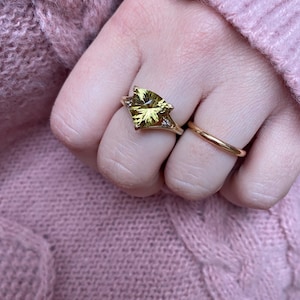 May include: A gold ring with a large, light green gemstone in a unique cut, flanked by small diamonds. A simple gold band ring is also visible. The rings are on a hand, with a pink sweater in the background.
