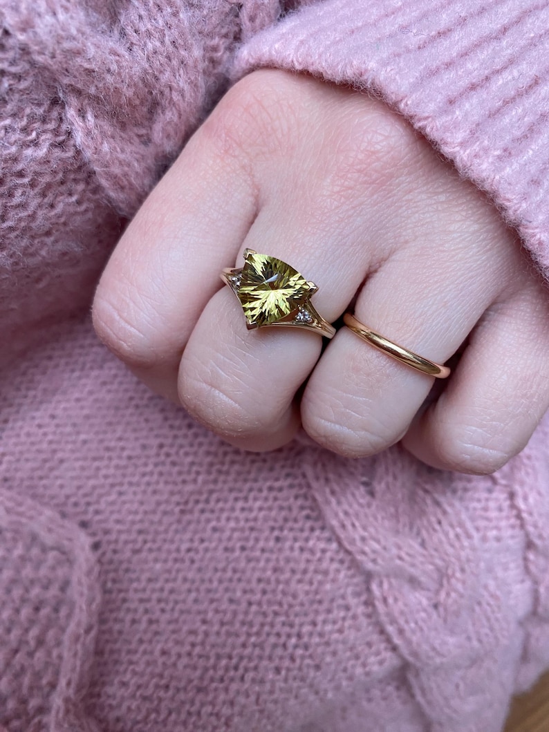 May include: A gold ring with a large, light green gemstone in a unique cut, flanked by small diamonds. A simple gold band ring is also visible. The rings are on a hand, with a pink sweater in the background.