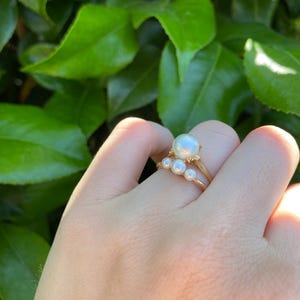 May include: A gold ring with a large, round, white pearl and three smaller pearls. The ring is held up against a background of green leaves.