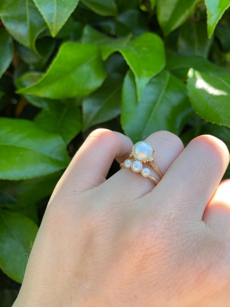May include: A gold ring with a large, round, white pearl and three smaller pearls. The ring is held up against a background of green leaves.