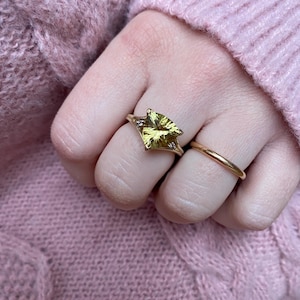 May include: A gold ring with a large, yellow-green gemstone in a triangular shape, set with small accent stones. A simple gold band ring is also visible. The rings are on a hand, with a pink sweater in the background.