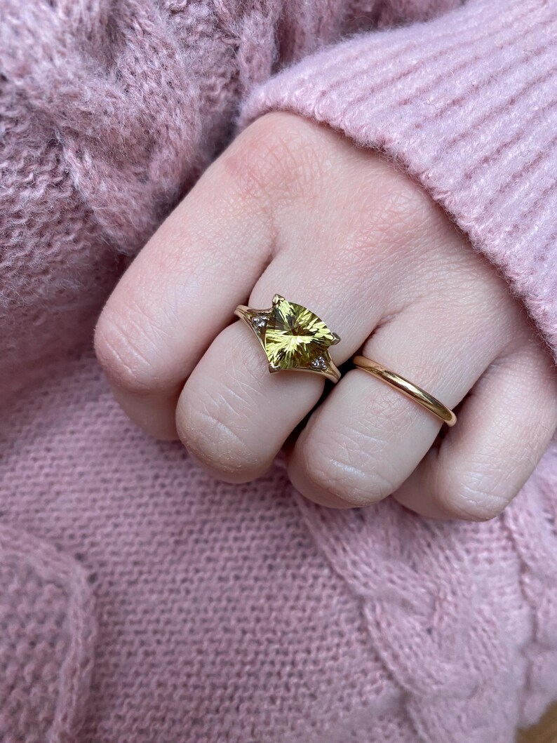 May include: A gold ring with a large, yellow-green gemstone in a triangular shape, set with small accent stones. A simple gold band ring is also visible. The rings are on a hand, with a pink sweater in the background.