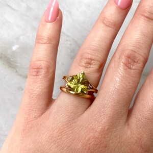 May include: A gold ring with a large, light green gemstone in a starburst design. The ring has a split shank and is adorned with small, clear accent stones. The ring is on a finger with pink nail polish.