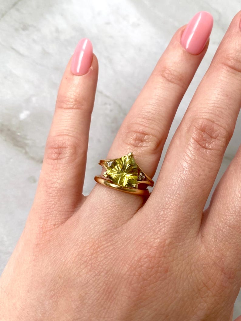 May include: A gold ring with a large, light green gemstone in a starburst design. The ring has a split shank and is adorned with small, clear accent stones. The ring is on a finger with pink nail polish.