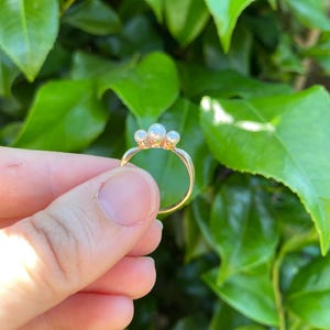 May include: A gold ring featuring three small, round pearls. The ring is held against a backdrop of green leaves, highlighting the delicate design and the contrast between the gold and the pearls. The ring's band is thin and elegant.