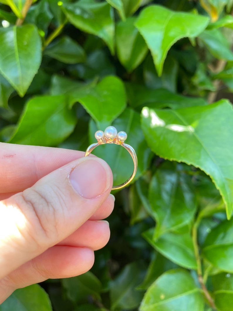 May include: A gold ring featuring three small, round pearls. The ring is held against a backdrop of green leaves, highlighting the delicate design and the contrast between the gold and the pearls. The ring's band is thin and elegant.