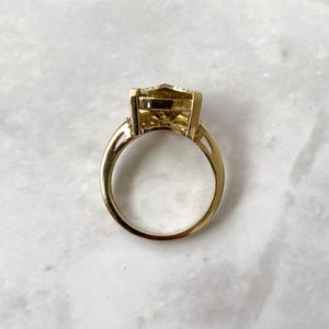 May include: A gold ring with a large, light-yellow gemstone. The ring has a classic design with a rectangular-cut stone and a delicate band. The gemstone is set in a gold bezel setting, and the band has a simple, elegant design.