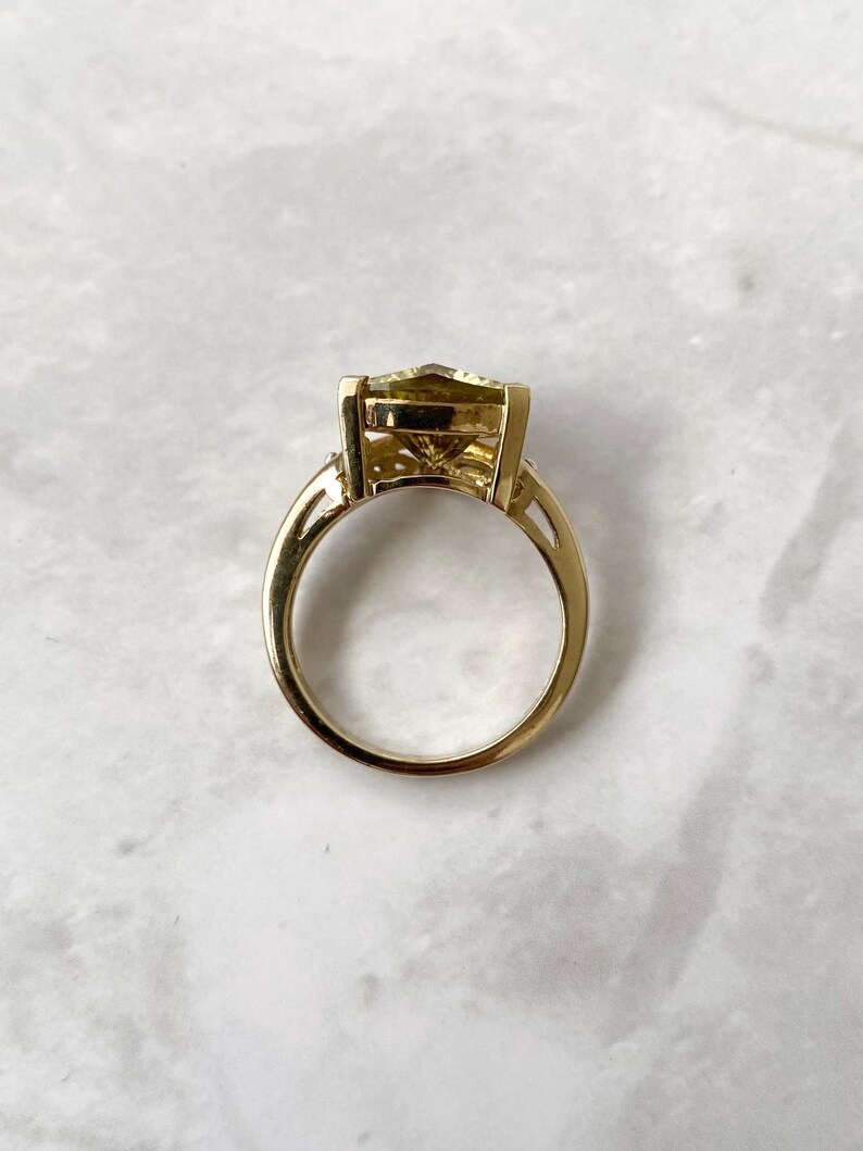 May include: A gold ring with a large, light-yellow gemstone. The ring has a classic design with a rectangular-cut stone and a delicate band. The gemstone is set in a gold bezel setting, and the band has a simple, elegant design.