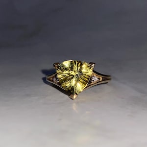 May include: A gold ring featuring a large, triangular, yellow gemstone. The ring has a delicate band with small accent stones on either side of the main stone. The gemstone has a faceted design, catching the light.