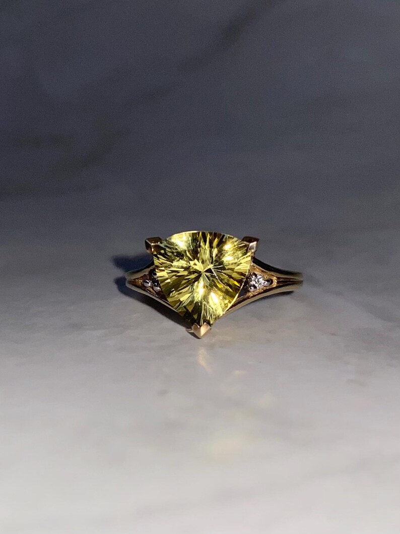 May include: A gold ring featuring a large, triangular, yellow gemstone. The ring has a delicate band with small accent stones on either side of the main stone. The gemstone has a faceted design, catching the light.