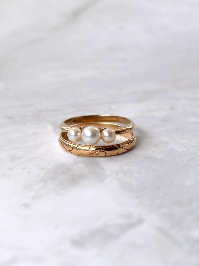 May include: Three gold rings are stacked on a white surface. The top ring has three white pearls. The middle ring is plain gold. The bottom ring has a floral design etched into the gold band.