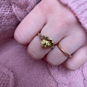 May include: A gold ring with a large, yellow-green gemstone in a leaf shape. A simple gold band ring is also visible. The rings are on a hand, with a pink sweater in the background.