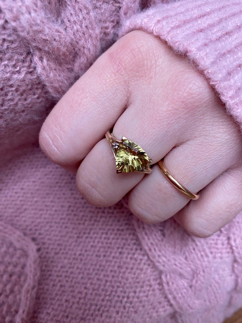 May include: A gold ring with a large, yellow-green gemstone in a leaf shape. A simple gold band ring is also visible. The rings are on a hand, with a pink sweater in the background.