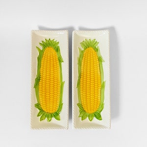 May include: Two white ceramic serving dishes with a raised design of corn on the cob. The corn is yellow with green husks.