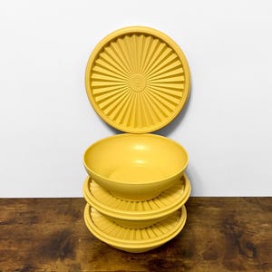 May include: A stack of three yellow plastic bowls with matching lids. The lids have a sunburst design. The bowls and lids are on a wooden surface with a white background. The bowls are a bright, solid yellow color.