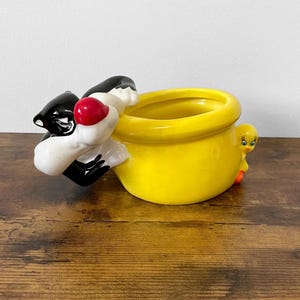 May include: A yellow ceramic planter featuring Sylvester the Cat and Tweety Bird. Sylvester is black and white with a red nose, clinging to the side. Tweety is yellow with orange feet. The planter is a bright yellow color.