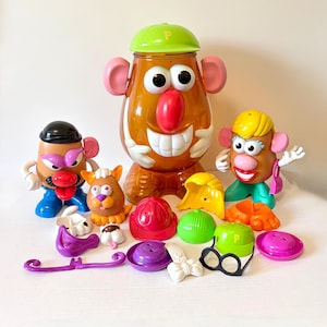 May include: A large translucent orange plastic Mr. Potato Head storage container with a green cap, accompanied by two smaller Mr. Potato Head figures, a small potato-shaped pet, and various plastic accessories including hats, shoes, and glasses.