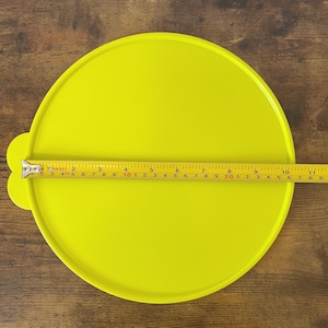 May include: A bright yellow, round plastic lid with a small handle. A yellow tape measure is laid across the lid, showing a diameter of approximately 10 inches. The lid is on a brown wooden surface.
