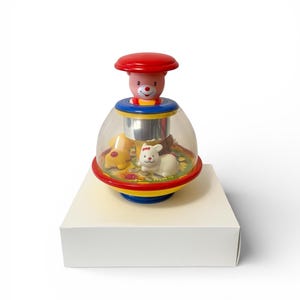 May include: A vintage baby toy with a red top and a pink bear face. The clear plastic dome contains a yellow dog and a white rabbit. The base is red, yellow, and blue. The toy sits on a white box.