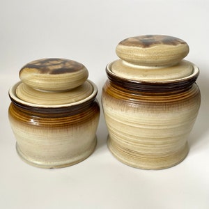 May include: Two ceramic jars with lids. The jars are beige with brown accents and a textured, swirling design. The lids are round and have a darker brown pattern on top. The jars are of different sizes.