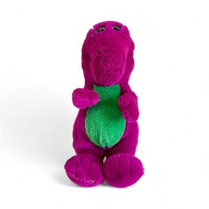 May include: A plush purple dinosaur toy with a green belly. The toy has a friendly expression with black eyes and is standing upright. The dinosaur's feet and hands are also purple, and the toy is set against a white background.